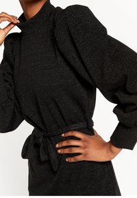 Mustapuku, jossa on korkea kaula, puffihihat ja teksturoitu kangas, jossa on hopeanvärisiä säikeitä. Sisältää sidottavan vyötäröyksityiskohdan.