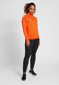 Top sportivo arancione con colletto alto, maniche lunghe e logo; abbinato a leggings neri con un motivo bianco sui lati e scarpe nere.