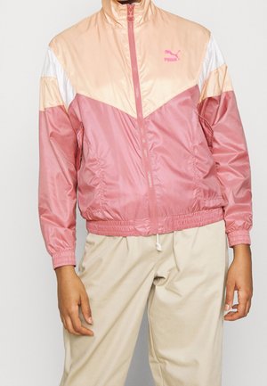 Personne portant une veste zippée Puma rose et pêche à manches longues, associée à un pantalon beige avec cordon de serrage, debout devant un fond uni.