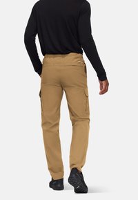 Mężczyzna w beżowych spodniach cargo, czarnej koszuli z długim rękawem i czarnych butach, odchodzi od jednego tła.