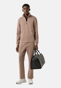 Jasnobrązowa bluza na zamek z długim rękawem i stójką, w zestawie z pasującymi spodniami z bocznymi szwami. Czarna skórzana torba sportowa. Białe trampki.