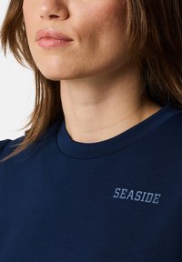 T-shirt in cotone navy con collo rotondo e piccola stampa "SEASIDE" in grigio. Il tessuto appare liscio con una vestibilità standard.
