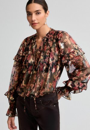 Blusa nera trasparente con motivo floreale in rosso, oro e verde. Maniche arricciate, scollatura con nodo e tessuto testurizzato. Indossata con pantaloni scuri.