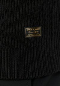 Sort ribbet strikket stof har en guld-tone label med teksten "JACK & JONES Superior Apparel" i nederste kant.