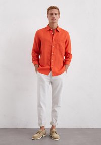 Orange linen button-up shirt with a chest pocket, paired with white rolled-up trousers and beige lace-up shoes. Plain background.