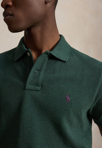 Muž nosi tmavozelené polo tričko s malým fialovým vyšívaným logom na ľavej hrudi, otočený doľava na jednofarebnom pozadí.