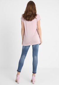 Kvinna som står med ryggen mot kameran, klädd i en ljusrosa kortärmad topp, blå smala jeans och rosa högklackade sandaler mot vit bakgrund.