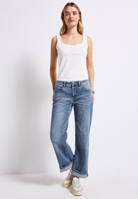 Top bianco leggero abbinato a jeans larghi azzurri con orlo cropped. Il modello indossa sneakers beige.