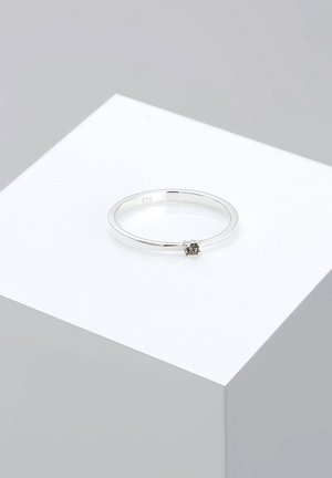 Silberner Ring mit dünnem Band, mit einem kleinen schwarzen Stein in der Mitte, auf einem weißen Sockel vor einem grauen Hintergrund präsentiert.
