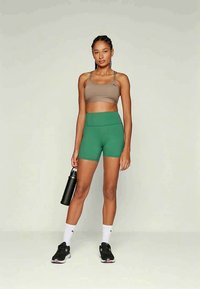 Szőke nő barna sportmelltartóban, zöld sortban, fehér zokniban és fekete sportcipőben, fekete vízpalackot tartva, világos háttér előtt áll.