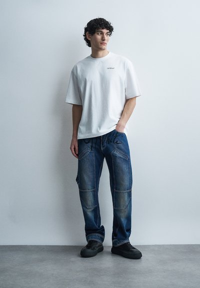 Jeune homme aux cheveux bouclés portant un t-shirt blanc surdimensionné, un pantalon cargo en denim bleu avec des détails de couture, et des baskets noires, debout à l'intérieur.