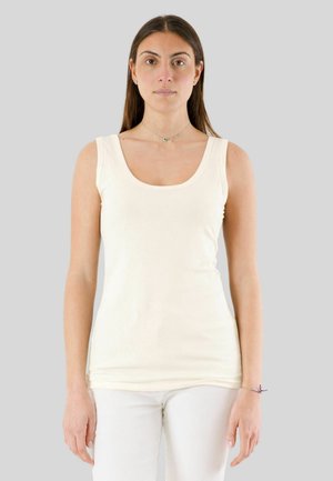 Giovane donna con lunghi capelli castani, che indossa un top bianco senza maniche e pantaloni bianchi, in piedi davanti a uno sfondo grigio uniforme.