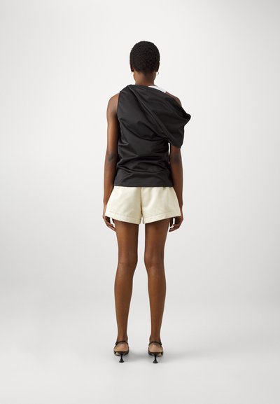 Top nero asimmetrico senza maniche, abbinato a pantaloni corti color crema. Il materiale sembra morbido, con una vestibilità ampia e un design drappeggiato.