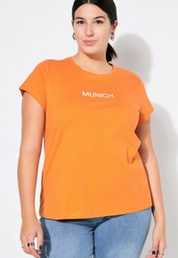 Orange Kurzarm-T-Shirt mit weißem "MÜNCHEN"-Text auf der Vorderseite. Weicher Baumwollstoff, lässige Passform, runder Halsausschnitt und subtile Seitenschlitze.