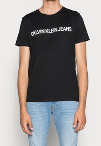 Man med en svart kortärmad T-shirt med "CALVIN KLEIN JEANS" i vita bokstäver, ihop med ljusblå jeans.