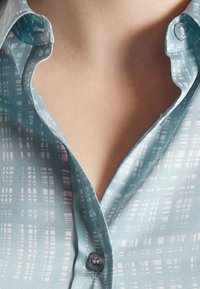 Chemise bleu clair à col, faite d'un tissu lisse avec un motif en grille en blanc et rose. Elle présente un bouton au col et une douce brillance.