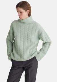 Minzgrüner Strickpullover mit hohem Kragen, mit Zopfmuster, lockerem Sitz und gerippten Bündchen. Kombiniert mit dunkler, maßgeschneiderter Hose.