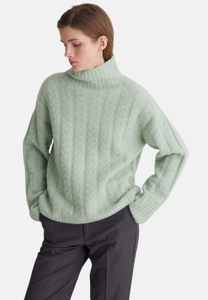 Suéter de punto verde menta con cuello alto, con patrón de trenzas, ajuste relajado y puños acanalados. Combinado con pantalones oscuros de corte entallado.