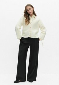 Kremowy, puszysty sweter kardigan z zapięciem na guziki, zestawiony z czarnymi spodniami o szerokich nogawkach i czarnymi butami wsuwanymi. Długie rękawy z wiązaniami.