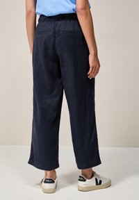 Osoba v volných námořnických zkrácených kalhotách, světle modrém topu a bílých teniskách Veja s černými akcenty, stojí na jednolitém pozadí.