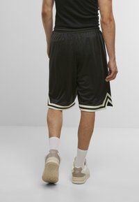 Black athletic shorts with side stripes in white, made of lightweight fabric. Worn with white socks and beige sneakers, viewed from the back.