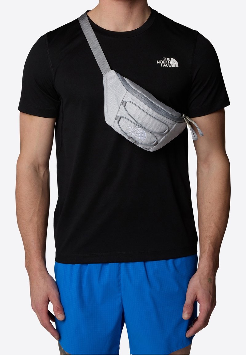 Homme portant un t-shirt noir The North Face, un short bleu et un sac banane gris à bandoulière avec le logo The North Face sur la poitrine et le sac.
