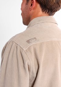 Camisa de pana beige con rayas verticales, que presenta un cuello clásico y una etiqueta en el hombro que muestra la marca "Oxbow."