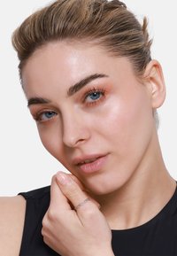 Primo piano di una persona con pelle chiara, capelli castano chiaro, occhi blu, che indossa un delicato anello d'argento. Pelle pulita con trucco sobrio e abbigliamento neutro.