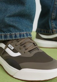 Brune syntetiske sneakers med hvite detaljer, teksturert stoff, flate lisser og gummisåle, sammen med blå denimjeans.