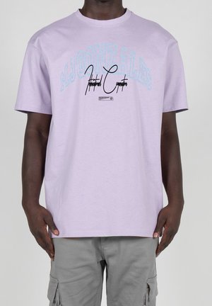 Mann trägt ein hellviolettes T-Shirt mit blauem und schwarzem Text "M.J. GONZALES" und "Industrial Corporation" sowie graue Cargohosen.
