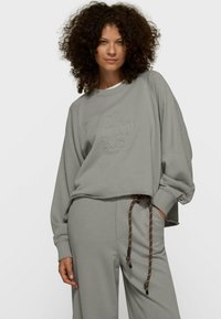 Sweat-shirt gris clair avec un col rond c ôtelé, épaules tombantes, et logo brodé. Associé à un pantalon ample assorti avec une taille à cordon de serrage.