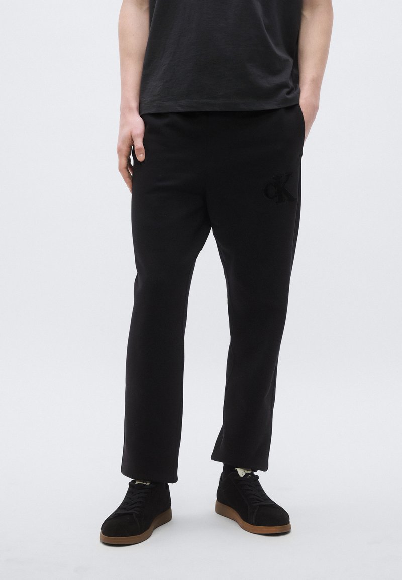 Μαύρο παντελόνι φόρμας με ελαστική μέση, διακοσμημένο με κεντημένο λογότυπο στον αριστερό μηρό, συνδυασμένο με μαύρα αθλητικά παπούτσια χαμηλού πάτου.