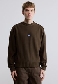 Pull marron en matière douce, avec un col et un ourlet côtelés. Une petite étiquette logo bleue est placée sur la poitrine.