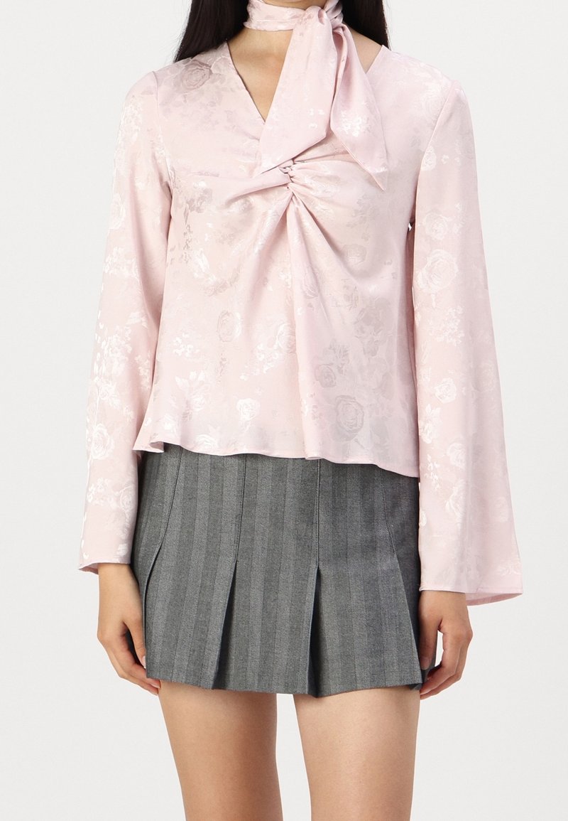Blusa di raso rosa con motivo floreale, dettaglio attorcigliato davanti e maniche a campana lunghe, abbinata a una gonna plissettata a righe grigie.