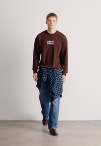 Καφέ φούτερ με λαιμόκοψη και λευκό λογότυπο, μπλε καρό πουκάμισο δεμένο στη μέση, μπλε τζιν, και καφέ δερμάτινα παπούτσια, περπατώντας σε ουδέτερο φόντο.