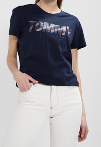 Marineblaue kurzärmlige T-Shirt aus Baumwolle mit einem grafischen "TOMMY"-Logo in mehrfarbigen Streifen. Tragen mit hellen Jeans.