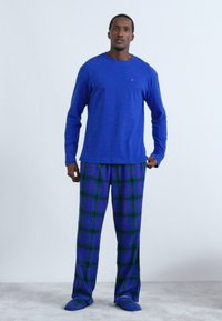 Modrá košeľa s dlhým rukávom, kockované nohavice v zelenej a fialovej farbe, zladené papuče, stojace na svetlošedej koberci na jednoduchom pozadí.