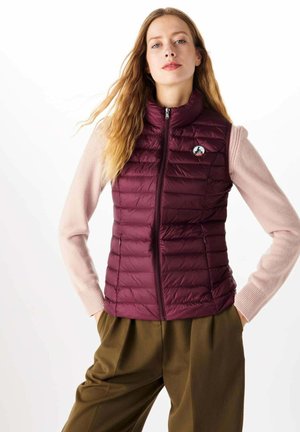 Doudoune sans manches bordeaux avec un design matelassé, un col montant et une fermeture éclair intégrale. Elle arbore un patch logo sur la poitrine et est portée par-dessus un pull rose clair.