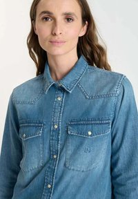 Kvinna med långt brunt hår som bär en knappad ljusblå denimskjorta med två framfickor med fälllock och tryckknappar.
