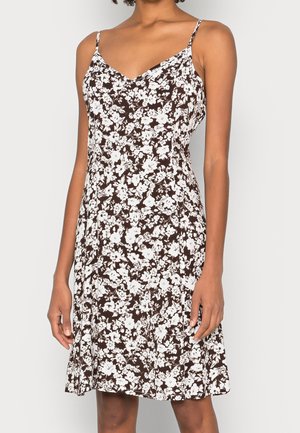Femme portant une robe marron sans manches avec un motif floral blanc et de fines bretelles, debout devant un fond clair uni.