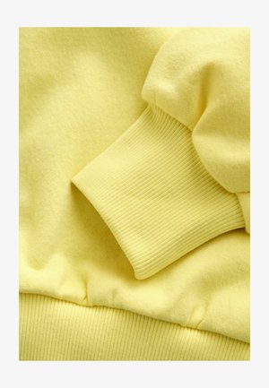 Żółta tkanina bluzy z miękkim, teksturowanym wykończeniem. Żebrowane mankiety i pas biodrowy, z bezszwowym designem dla gładkiego wyglądu.