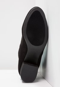Zapato de tacón alto de ante negro con suela de goma texturizada, que presenta un respaldo sólido y redondeado, y un acabado liso a lo largo del borde exterior.