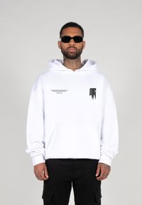White cotton hoodie with a front pocket, large hood, and black text details on the front. Worn with black cargo pants and sunglasses.