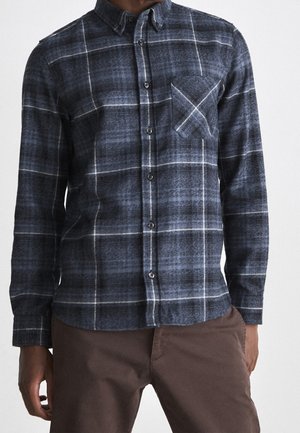 Homme portant une chemise à carreaux bleu foncé et gris à manches longues avec une poche poitrine et un pantalon marron.
