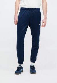 Tengerészkék atlétikai nadrág fehér oldalsávokkal, rugalmas derékrésszel és sima anyaggal, kék tornacipővel és fehér zoknival párosítva.