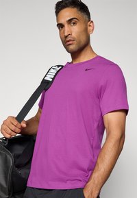Lilla Nike t-skjorte med rund hals og korte ermer. Har en liten svart logo på brystet og tekstil med struktur. Svart treningsbag i hånden.