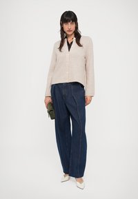 Beige gebreid vest met knopen aan de voorkant, gecombineerd met wijde donkere spijkerbroek met zichtbare stiksels. Casual witte schoenen met spitse neus.