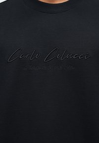 Μαύρο μπλουζάκι με στρογγυλή λαιμόκοψη, κεντημένο με το κείμενο "Carlo Colucci" σε καλλιγραφικό στυλ και το κείμενο "A Legacy Since 1978" με μικρότερα γράμματα από κάτω.