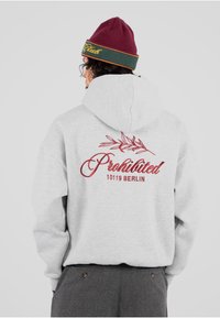 Sudadera gris con un gran texto bordado en rojo que dice "Prohibido" y un diseño de hojas en la parte de atrás. Se lleva con un gorro burdeos.