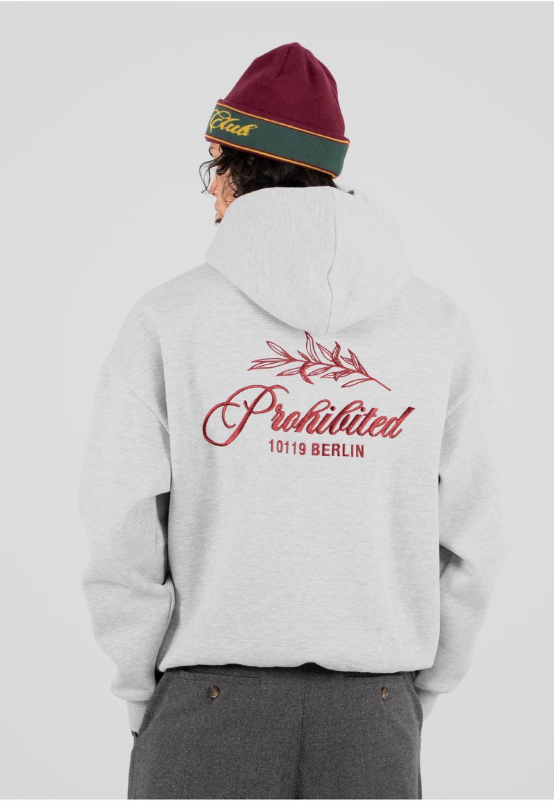 Sudadera gris con un gran texto bordado en rojo que dice "Prohibido" y un diseño de hojas en la parte de atrás. Se lleva con un gorro burdeos.